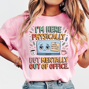Puede incluir: Camiseta rosa con el texto "I'M HERE PHYSICALLY BUT MENTALLY OUT OF OFFICE". El diseño incluye un portátil triste, tazas de café y clips. La camiseta es de cuello redondo.