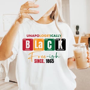 May include: White t-shirt with the words "UNAPOLOGETICALLY BLACK Free-ish SINCE 1865" in a colorful design. The word "BLACK" is spelled out using elements from the periodic table. The shirt is worn by a person holding a phone and a glass of iced coffee.