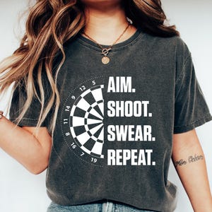 May include: Charcoal grey t-shirt featuring a dartboard design and the words "AIM. SHOOT. SWEAR. REPEAT." The shirt is worn with blue jeans and white trainers. A necklace and bracelets are also visible.