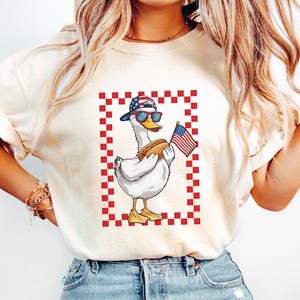 Op de afbeelding: Een wit T-shirt met een rood-wit-blauwe geruite rand rond een cartoonafbeelding van een eend die een rood-wit-blauwe baseballpet, zonnebril draagt en een hotdog en een Amerikaanse vlag vasthoudt.
