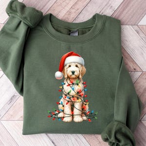 May include: An olive green sweatshirt featuring a graphic of a golden retriever puppy wearing a Santa hat and wrapped in Christmas lights. The puppy is sitting, and the lights are multi-colored. The sweatshirt has long sleeves and a crew neck.