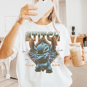 May include: White t-shirt featuring a graphic of Stitch from Lilo & Stitch. The design includes the word "Stitch" in a retro font, Stitch's image, and the numbers "626" and "CKE". The shirt is a casual, oversized fit.