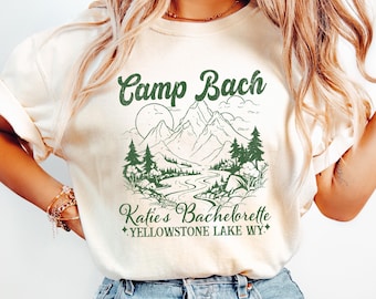 May include: Cream-colored t-shirt with a green graphic of a mountain landscape and the text "Camp Bach" and "Katie's Bachelorette Yellowstone Lake WY". The shirt is paired with blue jeans.