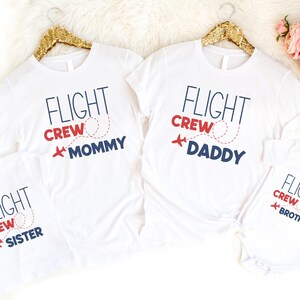 以下が含まれることがあります： 白地に「FLIGHT CREW」の文字がネイビー、赤、白でプリントされたTシャツ。シャツには「Mommy」、「Daddy」、「Sister」、「Brother」と書かれています。赤い飛行機のグラフィックと点線も特徴です。