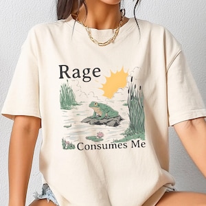 May include: Cream-colored t-shirt featuring a graphic of a frog on a rock in a pond, with the text "Rage Consumes Me." The design includes a sun, reeds, and lily pads. The shirt has short sleeves.