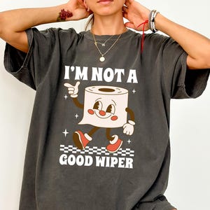 May include: Dark gray t-shirt with the text "I'M NOT A GOOD WIPER" and a cartoon toilet paper roll character. The character has a smiling face, arms, legs, and red shoes. The shirt is a relaxed fit.