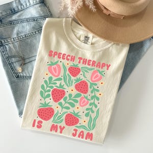 May include: Cream-colored t-shirt with the text "SPEECH THERAPY IS MY JAM" in pink. The shirt features a strawberry design with green leaves. The shirt is paired with light blue jeans, a gold necklace, and a tan hat.