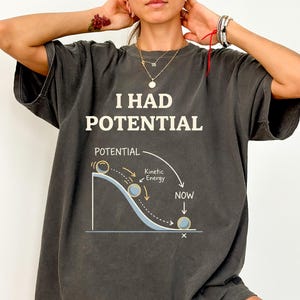 May include: Dark gray t-shirt with the text "I HAD POTENTIAL" above a diagram illustrating potential and kinetic energy. The diagram shows a ball rolling down a curved path, with labels indicating "Potential," "Kinetic Energy," and "NOW."