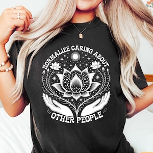 Può includere: T-shirt nero con un design grafico bianco raffigurante un fiore di loto, mani e la scritta "NORMALIZE CARING ABOUT OTHER PEOPLE". Il design include un sole e delle stelle.