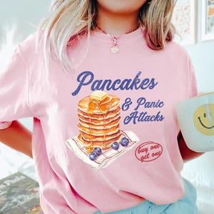 May include: Pale pink t-shirt featuring the text "Pancakes & Panic Attacks" and a pancake illustration with butter, syrup, and blueberries. A white mug with a yellow smiley face is visible.
