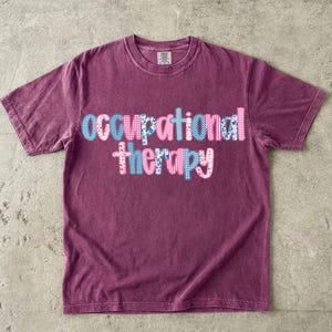 Könnte beinhalten: Ein kastanienbraunes Kurzarm-T-Shirt mit dem Schriftzug occupational therapy auf der Brust. Die Buchstaben sind mit verschiedenen Mustern gefüllt, darunter blau-weißes Vichy-Karo, rosa Streifen sowie florale Designs in verschiedenen Rosa- und Blautönen.