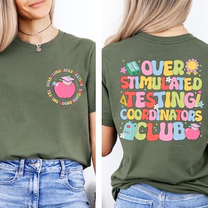 Può includere: T-shirt verde oliva con la scritta "OVER STIMULATED TESTING COORDINATORS CLUB" in lettere colorate in stile retrò. La parte anteriore presenta una grafica di una mela con cappello di laurea. La maglietta è abbinata a jeans blu.