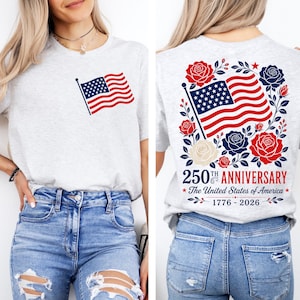 Puede incluir: Camiseta gris claro con la bandera estadounidense en la parte delantera y trasera. La parte trasera de la camiseta muestra el texto "250th Anniversary The United States of America 1776-2026" rodeado de elementos florales rojos, blancos y azules.