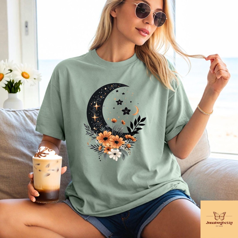 May include: Sage green t-shirt featuring a crescent moon design filled with stars and accented with orange and white flowers. The graphic tee has a relaxed fit and short sleeves, perfect for casual wear.