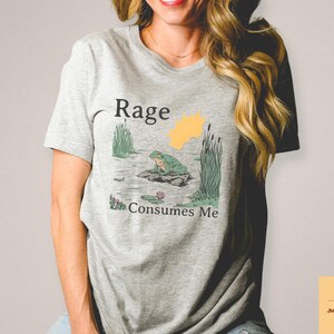 May include: A light gray t-shirt featuring a graphic of a frog on a rock in a pond, with the words "Rage Consumes Me." The design includes a yellow sun and green reeds. The shirt is made of a soft material.