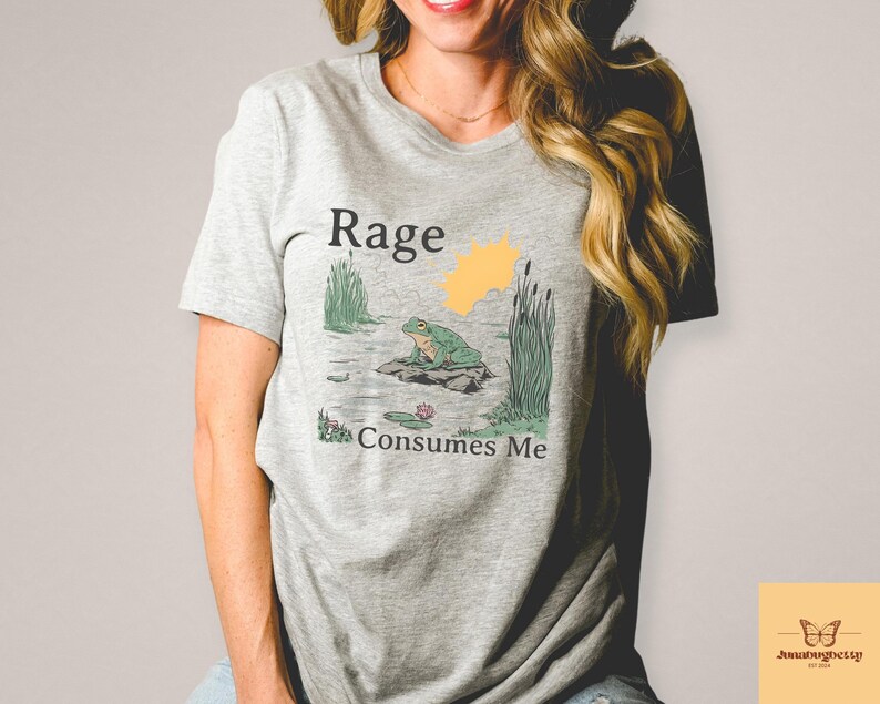 May include: A light gray t-shirt featuring a graphic of a frog on a rock in a pond, with the words "Rage Consumes Me." The design includes a yellow sun and green reeds. The shirt is made of a soft material.