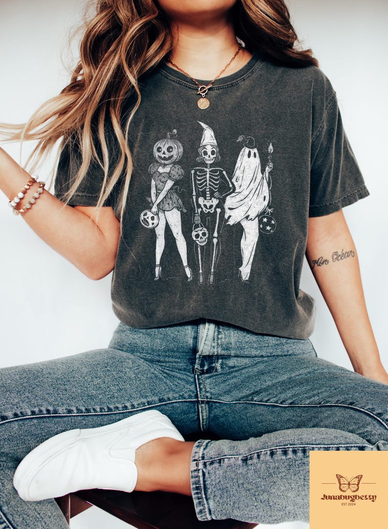 May include: Dark gray t-shirt featuring a Halloween-themed graphic. The design includes a pumpkin-headed figure, a skeleton, and a ghost, each holding a trick-or-treat bag. The shirt is worn with blue jeans and white sneakers.