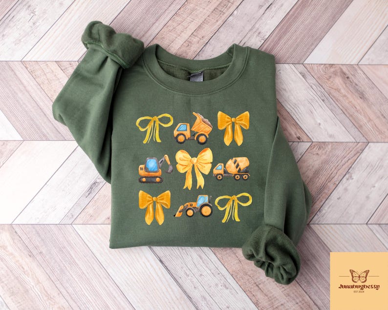 May include: An olive green sweatshirt with a watercolour design of construction vehicles and yellow bows. The design includes a dump truck, excavator, cement mixer, and tractor. The sweatshirt has long sleeves and a crew neck.