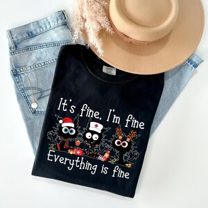 Może przedstawiać: Czarny t-shirt z napisem "It's fine, I'm fine, Everything is fine" i grafiką trzech kresk&oacute;wkowych kot&oacute;w. Koszulka jest połączona z jasnoniebieskimi dżinsami i beżowym kapeluszem.