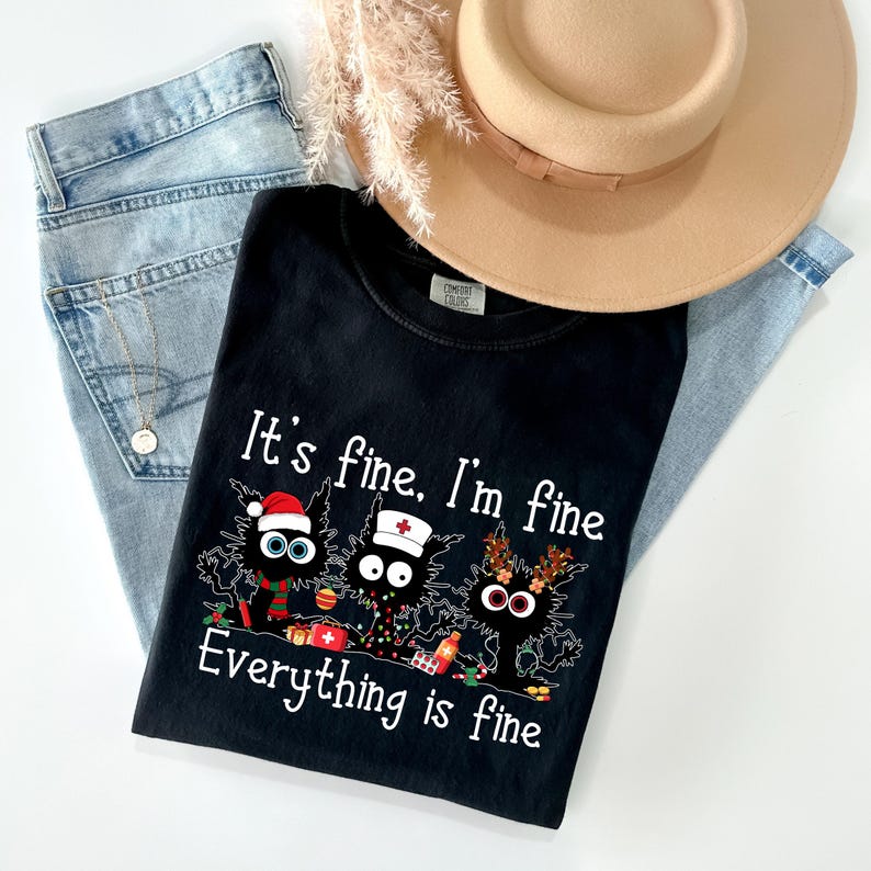 Może przedstawiać: Czarny t-shirt z napisem "It's fine, I'm fine, Everything is fine" i grafiką trzech kresk&oacute;wkowych kot&oacute;w. Koszulka jest połączona z jasnoniebieskimi dżinsami i beżowym kapeluszem.