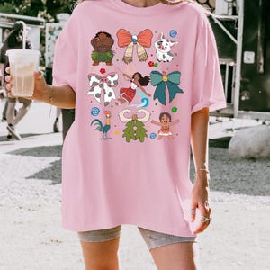 May include: Pink t-shirt featuring colorful cartoon illustrations of characters and bows. The design includes a variety of characters and bows in different colors and styles. The shirt is a casual, oversized fit.