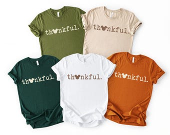Può includere: Cinque magliette a maniche corte con girocollo in verde oliva, beige, verde scuro, bianco e arancione bruciato. Ogni maglietta presenta la scritta "thankful." stampata con un carattere semplice, con una sagoma di testa di topo al posto della "a".