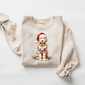May include: A cream-colored sweatshirt featuring a watercolor illustration of a golden doodle wearing a Santa hat and wrapped in Christmas lights. The dog is the central focus, with colorful lights adding festive detail.
