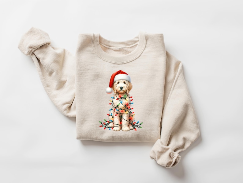 May include: A cream-colored sweatshirt featuring a watercolor illustration of a golden doodle wearing a Santa hat and wrapped in Christmas lights. The dog is the central focus, with colorful lights adding festive detail.
