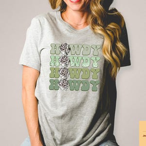 May include: A gray t-shirt with a retro-style graphic design featuring the word "Howdy" repeated four times in a gradient of green and brown colors. Each "Howdy" is decorated with a stylized green clover.