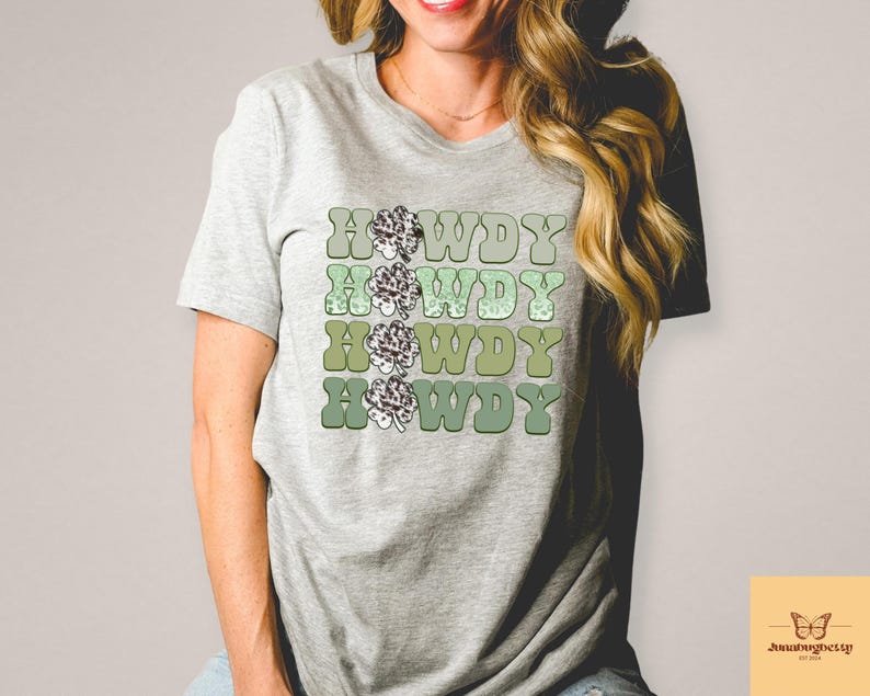 May include: A gray t-shirt with a retro-style graphic design featuring the word "Howdy" repeated four times in a gradient of green and brown colors. Each "Howdy" is decorated with a stylized green clover.
