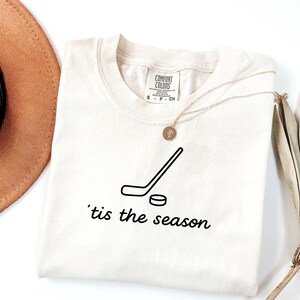 May include: Cream-colored Comfort Colors t-shirt featuring a black hockey stick and puck graphic. The text "'tis the season" is printed below the graphic. A gold necklace with a pendant is draped on the shirt.