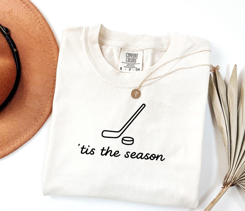 May include: Cream-colored Comfort Colors t-shirt featuring a black hockey stick and puck graphic. The text "'tis the season" is printed below the graphic. A gold necklace with a pendant is draped on the shirt.
