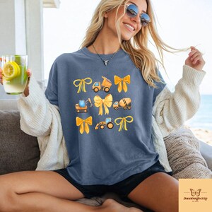 May include: A blue t-shirt with a design of construction vehicles and yellow bows. The illustrations include a dump truck, excavator, cement mixer, and a front-end loader. The shirt has a relaxed fit.