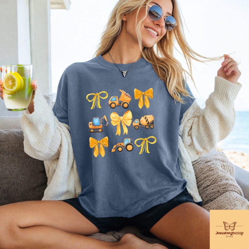 May include: A blue t-shirt with a design of construction vehicles and yellow bows. The illustrations include a dump truck, excavator, cement mixer, and a front-end loader. The shirt has a relaxed fit.