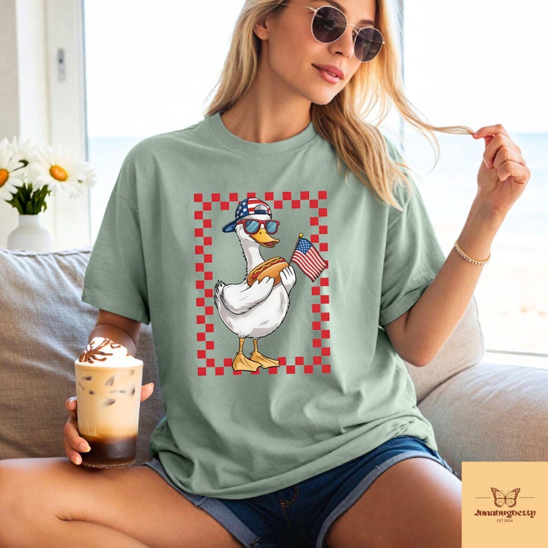 Op de afbeelding: Een groen T-shirt met een cartoon eend die een rood-wit-blauwe baseballpet draagt en een hotdog en een Amerikaanse vlag vasthoudt. De eend bevindt zich in een rood-wit geruit kader.