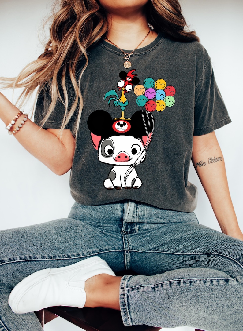 May include: Dark gray t-shirt featuring a cartoon pig wearing Mickey Mouse ears and holding a balloon bouquet. A cartoon rooster sits atop the pig's head. The shirt is a comfortable, casual style.