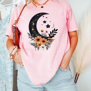 May include: Pink t-shirt featuring a celestial design with a crescent moon, stars, and floral accents. The graphic is in black, gold, and white. The shirt is a soft pink color and is made of a comfortable material.