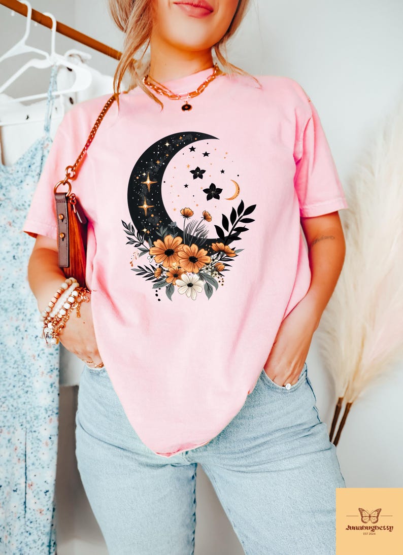 May include: Pink t-shirt featuring a celestial design with a crescent moon, stars, and floral accents. The graphic is in black, gold, and white. The shirt is a soft pink color and is made of a comfortable material.