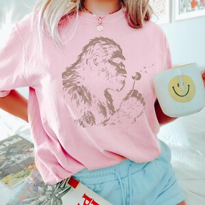Puede incluir: Sudadera rosa claro con un gr&aacute;fico marr&oacute;n de un bigfoot soplando un diente de le&oacute;n. La persona sostiene una taza blanca con una carita sonriente amarilla. La persona lleva pantalones cortos azul claro.