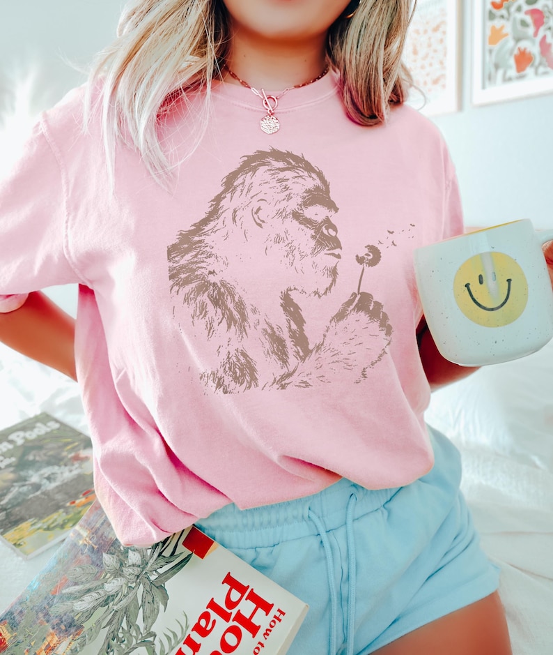 Puede incluir: Sudadera rosa claro con un gr&aacute;fico marr&oacute;n de un bigfoot soplando un diente de le&oacute;n. La persona sostiene una taza blanca con una carita sonriente amarilla. La persona lleva pantalones cortos azul claro.