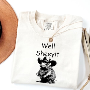 May include: Cream-coloured t-shirt featuring the text "Well Sheeeyit" and a black and white illustration of an armadillo wearing a cowboy hat. A gold necklace and a brown hat are also visible.