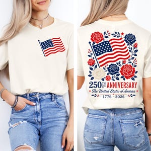 Puede incluir: Camiseta color crema con una peque&ntilde;a bandera estadounidense en la parte delantera y un dise&ntilde;o m&aacute;s grande en la parte trasera. El dise&ntilde;o trasero incluye una bandera ondeando, rosas rojas y azules, y el texto "250th Anniversary The United States of America 1776-2026."