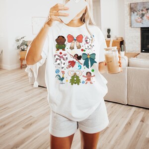 May include: White t-shirt featuring colorful cartoon character bows. The bows include a variety of designs, such as a red bow with gold accents, a green bow with a flower, and a blue bow with a gold accent.