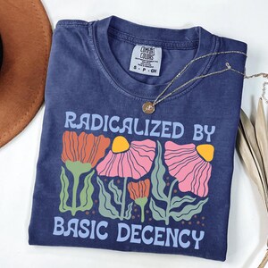 以下が含まれることがあります： 「RADICALIZED BY BASIC DECENCY」の文字とオレンジ、ピンク、黄色の花柄がプリントされたネイビーブルーのTシャツ。ペンダント付きのゴールドネックレスがシャツの上にかけられています。