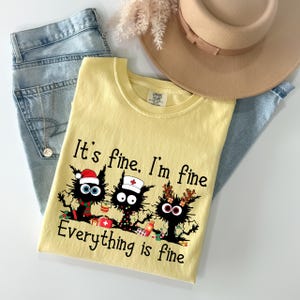 Może przedstawiać: Jasnoż&oacute;łta koszulka z napisem "It's fine. I'm fine. Everything is fine" i grafiką trzech czarnych kot&oacute;w. Koszulka jest zestawiona z jasnoniebieskimi dżinsami i beżowym kapeluszem.