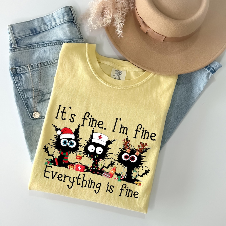 Może przedstawiać: Jasnoż&oacute;łta koszulka z napisem "It's fine. I'm fine. Everything is fine" i grafiką trzech czarnych kot&oacute;w. Koszulka jest zestawiona z jasnoniebieskimi dżinsami i beżowym kapeluszem.