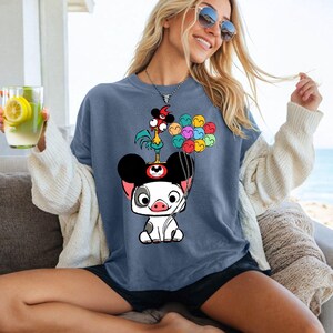 May include: A blue t-shirt featuring a cartoon pig wearing a Mickey Mouse hat, with a rooster and colorful balloons. The shirt is a relaxed fit, with short sleeves. The image is a graphic design.