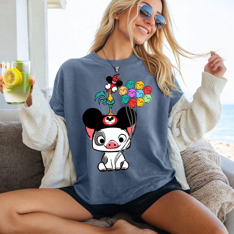 May include: A blue t-shirt featuring a cartoon pig wearing a Mickey Mouse hat, with a rooster and colorful balloons. The shirt is a relaxed fit, with short sleeves. The image is a graphic design.