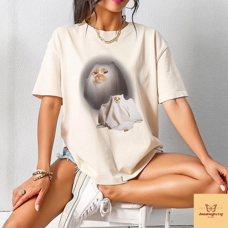 以下が含まれることがあります： クリーム色のTシャツ。猫をモチーフにしたデザインで、オレンジと白の毛並みのふわふわした猫と、幽霊に扮した2匹目の猫が描かれています。ゆったりとしたフィット感のシャツです。