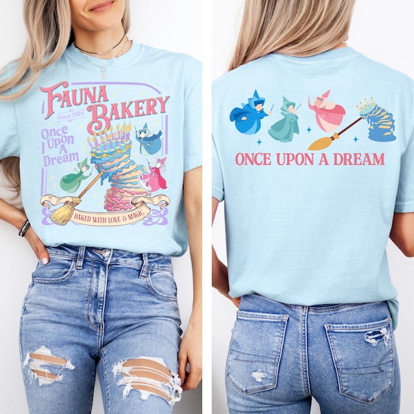 May include: A light blue t-shirt featuring a graphic on the front and back. The front displays a multi-tiered cake, three fairies, a broom, and text reading Fauna Bakery, Once Upon A Dream, and Baked With Love & Magic. The back shows three fairies.